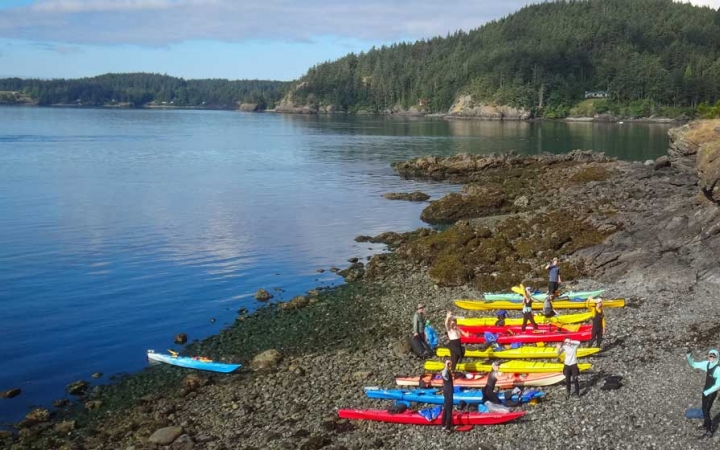 A row of colorful kayaks rest on the beach beside blue water., while people stand near them. There is a tree covered hill in the background. 